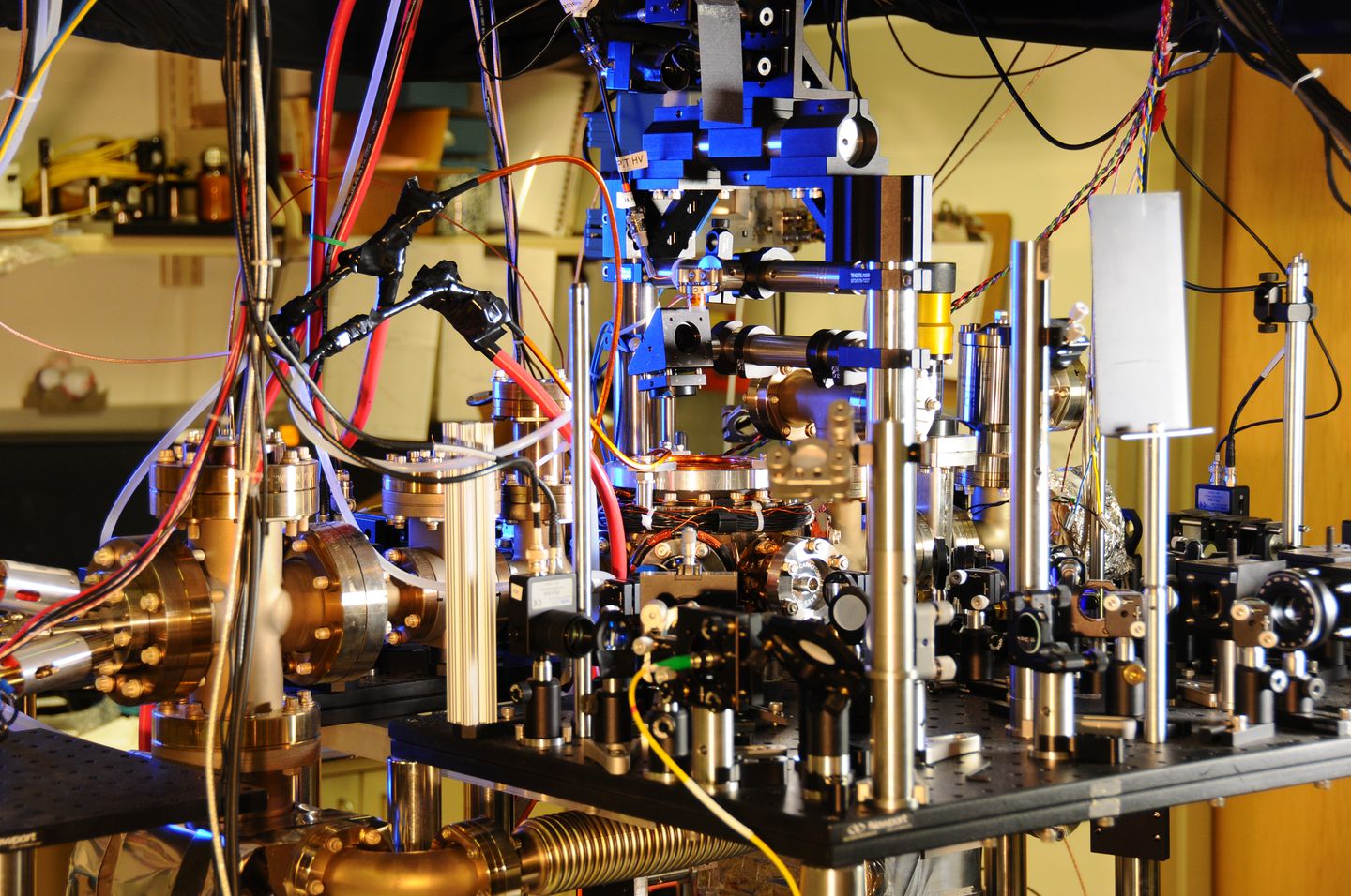 Lab bench at NIST with a metal oven on the left, a vacuum chamber in the center, and laser optics and a yellow optical fiber in front.
